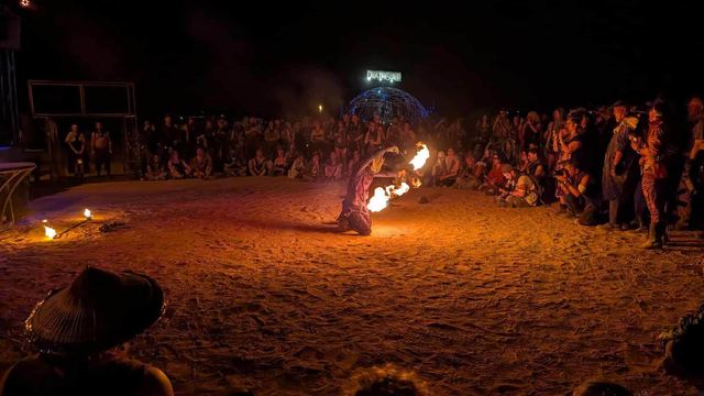 A fire performer kneels in the sand, arms spread, flames blooming from both hands before a night crowd.