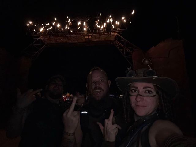 Three members at night beneath the gate, the WASTELAND letters lit by open flame high above.