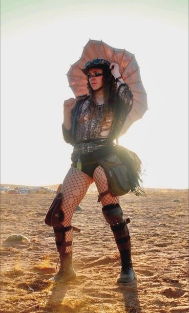 Sweet Tooth poses in the desert backlit by the sun, holding a tattered parasol, in goggles, fishnets, and armored boots.