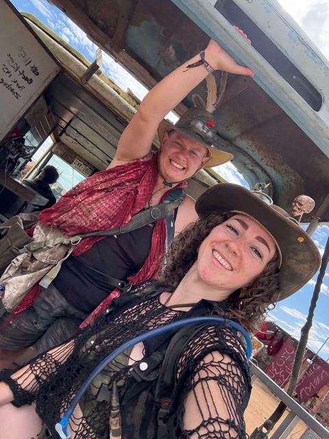 Two smiling members take a selfie in the shade beneath a rusted truck bed, blue sky behind.