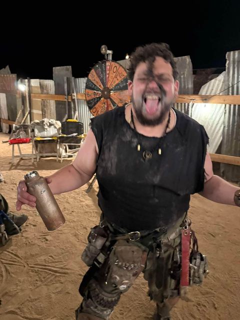Brisket at night, tongue out and arms spread, holding a battered steel bottle, a barter wheel of fortune behind him.