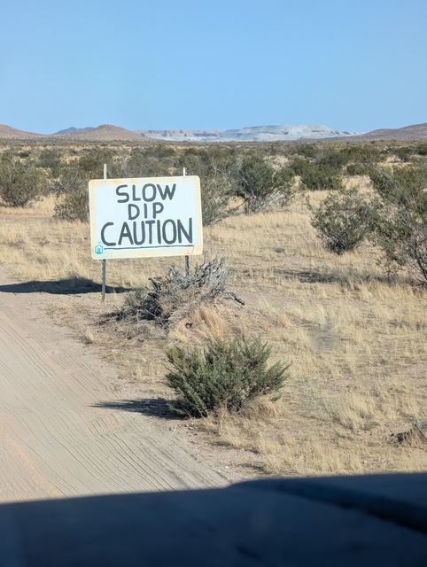 A hand-painted SLOW DIP CAUTION sign beside a dirt road through dry Mojave scrub.
