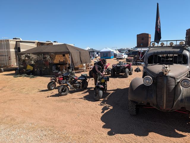 A black war car beside parked quads and motorcycles at a dusty campsite.