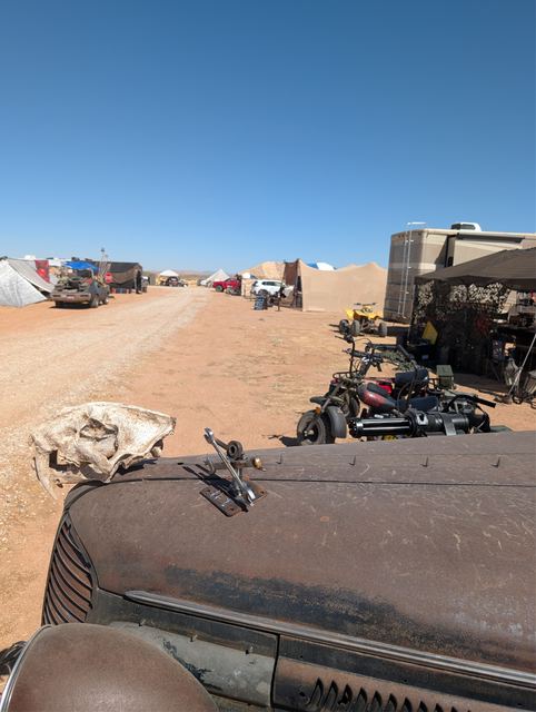 View over the war car's hood and skull ornament down a camp lane of tents and bikes.