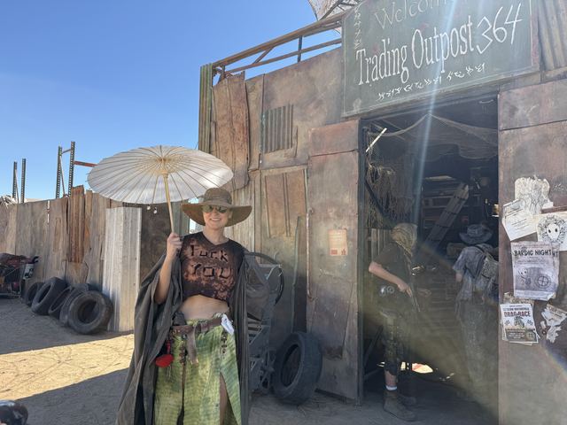 A wastelander with a white parasol and wide-brimmed hat poses in front of the Trading Outpost 364 shack.