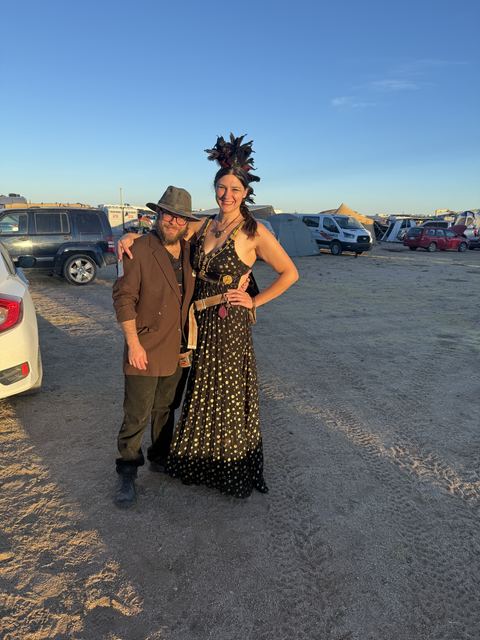 A couple dressed for evening — a brown coat and fedora, a sequined black gown and feather headdress — at dusk.
