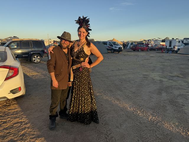 The same couple posing between parked cars at golden hour.