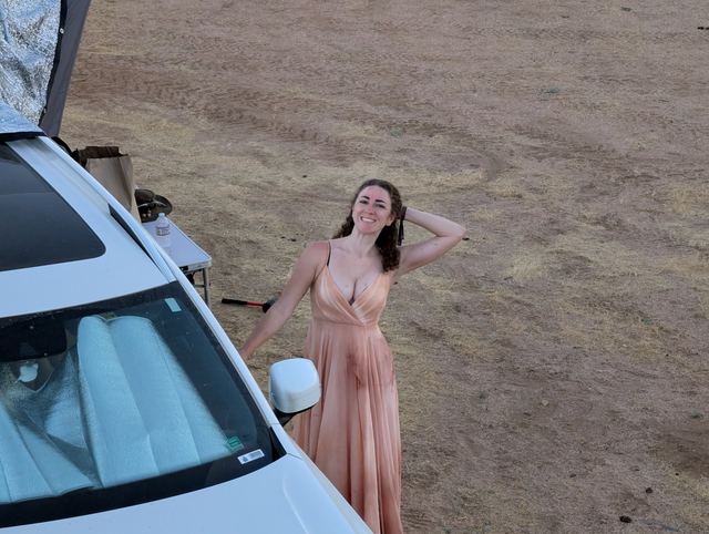 A woman in a long peach gown poses beside a white car in the desert.
