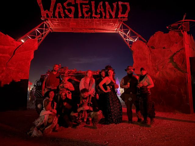 A group of costumed wastelanders posing under the red-lit Wasteland gate at night.