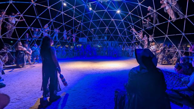 Two hooded attendants watch from the dome floor in blue light.