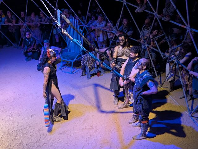 A woman in black with a feathered mohawk faces two fighters in the dome.
