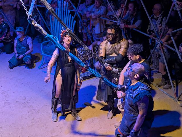 The feather-mohawked announcer and fighters gathered beneath the rigging.