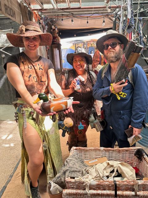 Three wastelanders in wide hats smile behind a crate of trade goods at a market stall.