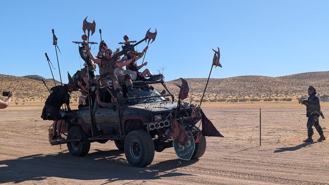 A spiked war truck bristling with axes and riders drives across the desert.