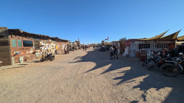 A long dusty street of shacks with motorcycles parked along it.