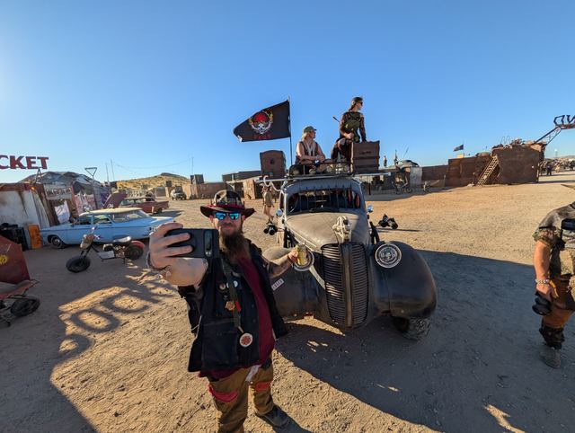 Grandpa in a red-banded hat takes a selfie in front of the war car, a flag flying behind.