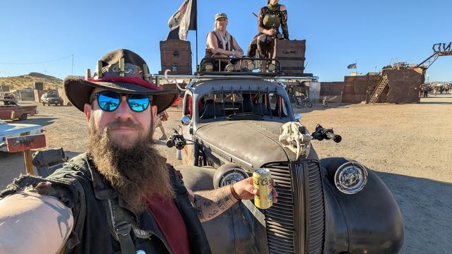 Selfie of Grandpa in mirrored sunglasses holding a drink, the skull-nosed war car and two riders behind.