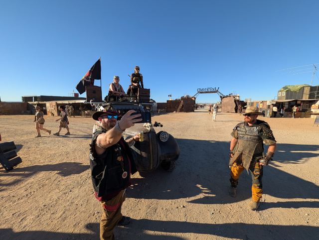 Grandpa snaps a selfie as another wastelander strolls up beside the war car.