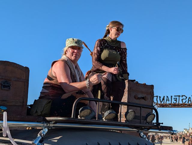 Two wastelanders sit on the war car's roof rack against a blue sky.