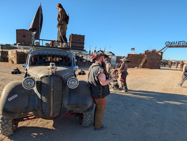 A wastelander stands on the war car roof, Grandpa leaning on the hood.