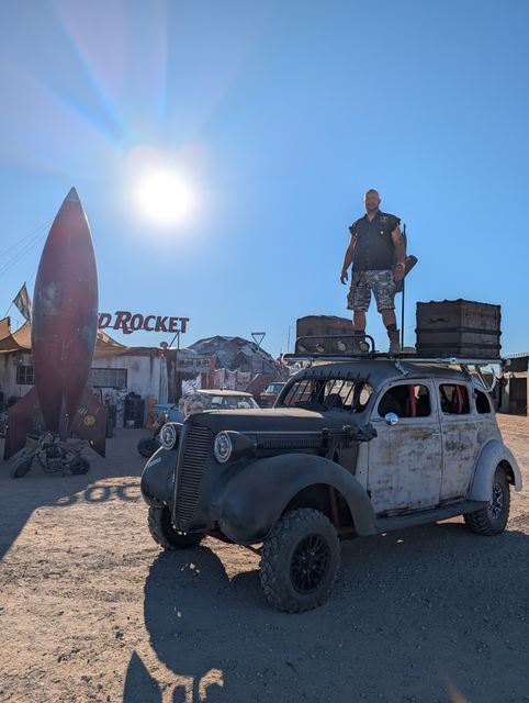 A wastelander stands on the war car's roof by the Red Rocket, the sun blazing behind.