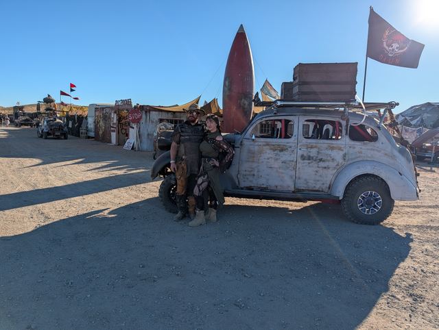 Two wastelanders pose by the war car under its black flag.