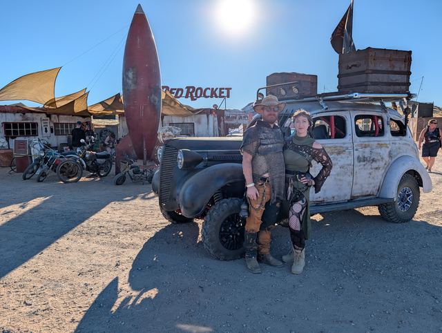 The couple in front of the war car, sun high over the rocket.