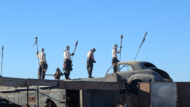 Four wastelanders with spears stand on a rooftop against the sky.