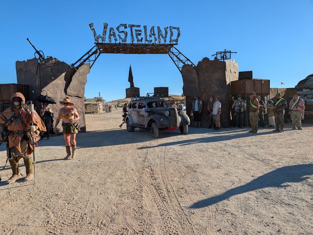 The war car under the Wasteland sign, wastelanders gathered around it.