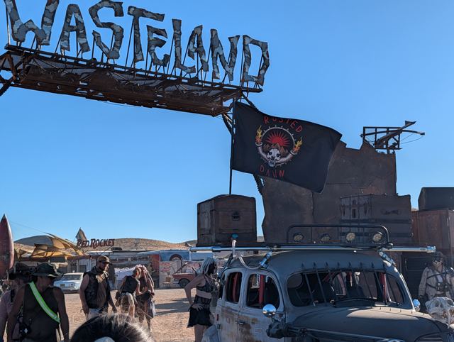 The Rusted Dawn flag flying from the war car beneath the WASTELAND sign.