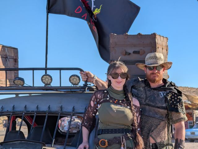 A woman in olive and a bearded man in a straw hat pose by the war car, the tribe flag above.