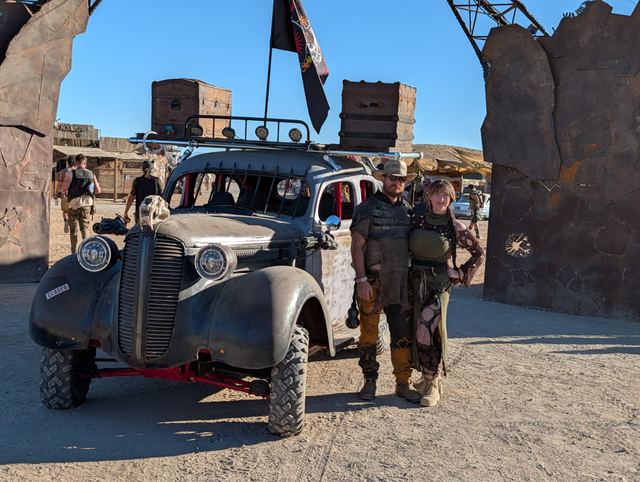 A couple in leather and chainmail poses by the war car under the gate.