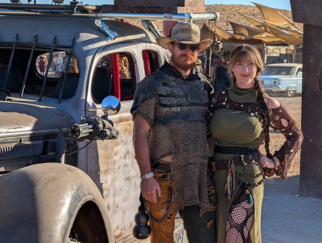 A man in chainmail and a woman in olive pose against the war car's side.