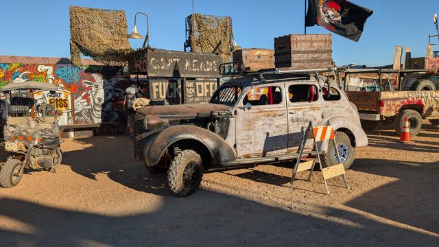 The war car parked at a camp frontage beside spray-painted signs.