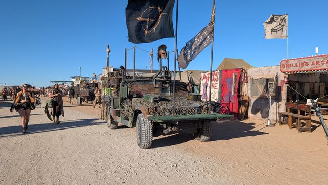 A military truck flying flags rolls down a camp street.