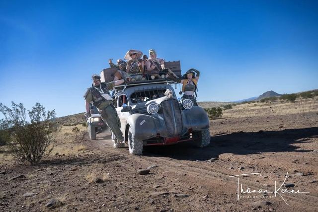 The war car crawling over desert scrub with riders on top. Photographer's watermark in the corner.