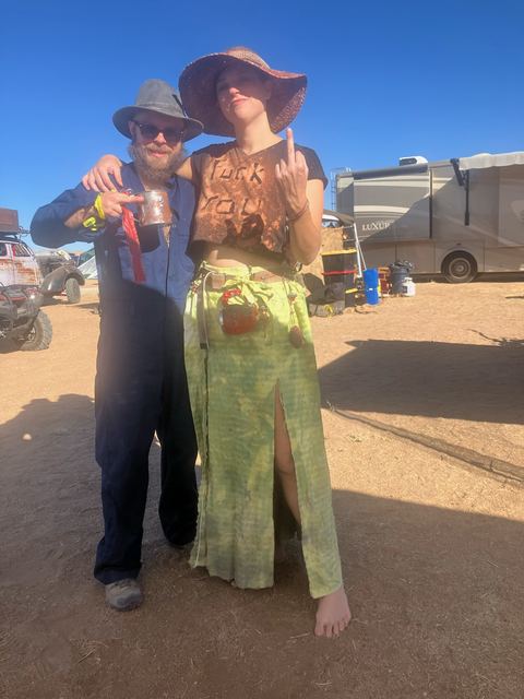 A man in a hat and goggles and a woman in a wide sun hat and green skirt pose at camp.