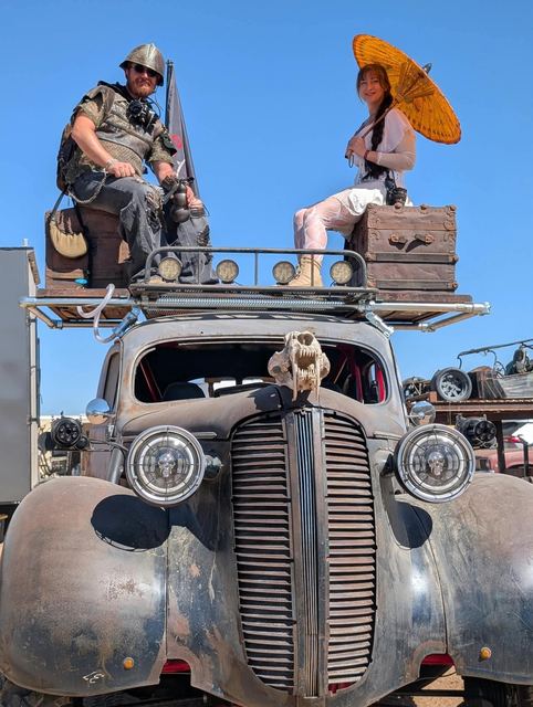 Two riders sit on the war car's roof, one with an orange parasol, the skull ornament on the grille.